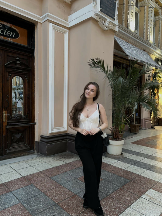 Street style photo, Fashion look, Casual-chic outfit, White corset top, Black wide-leg pants, Elegant jewelry, Designer bag, European architecture background, Evening light, Summer/Spring collection.